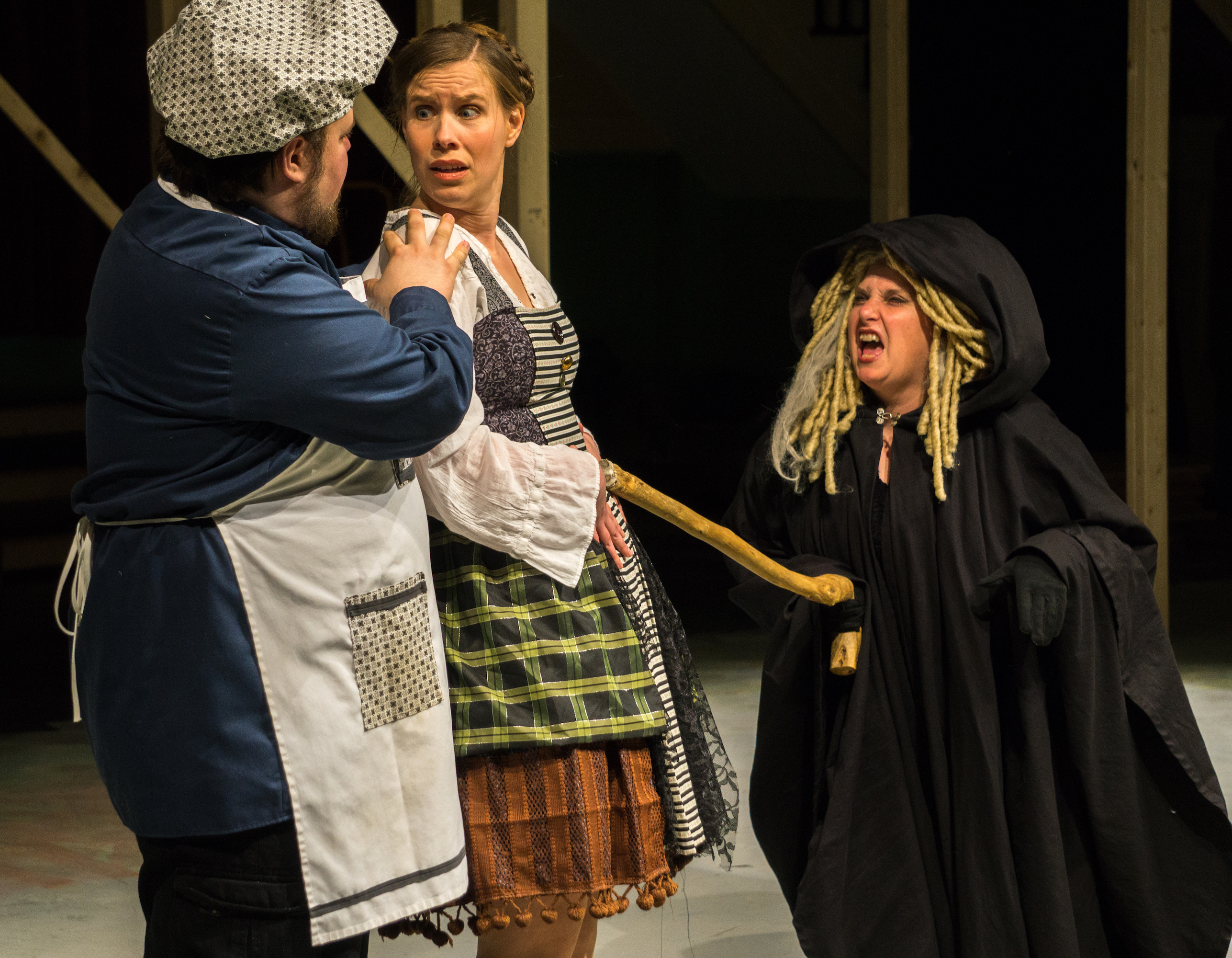 The Witch (cloaked in black) threatens The Bakers Wife (Carolyn Wesley in multi-colored, short skirted peasant outfit) by lunging at her with her magic cane, while the Baker (Nick Wheeler, in polka dotted baking cap & matching apron) tries to protect his wife